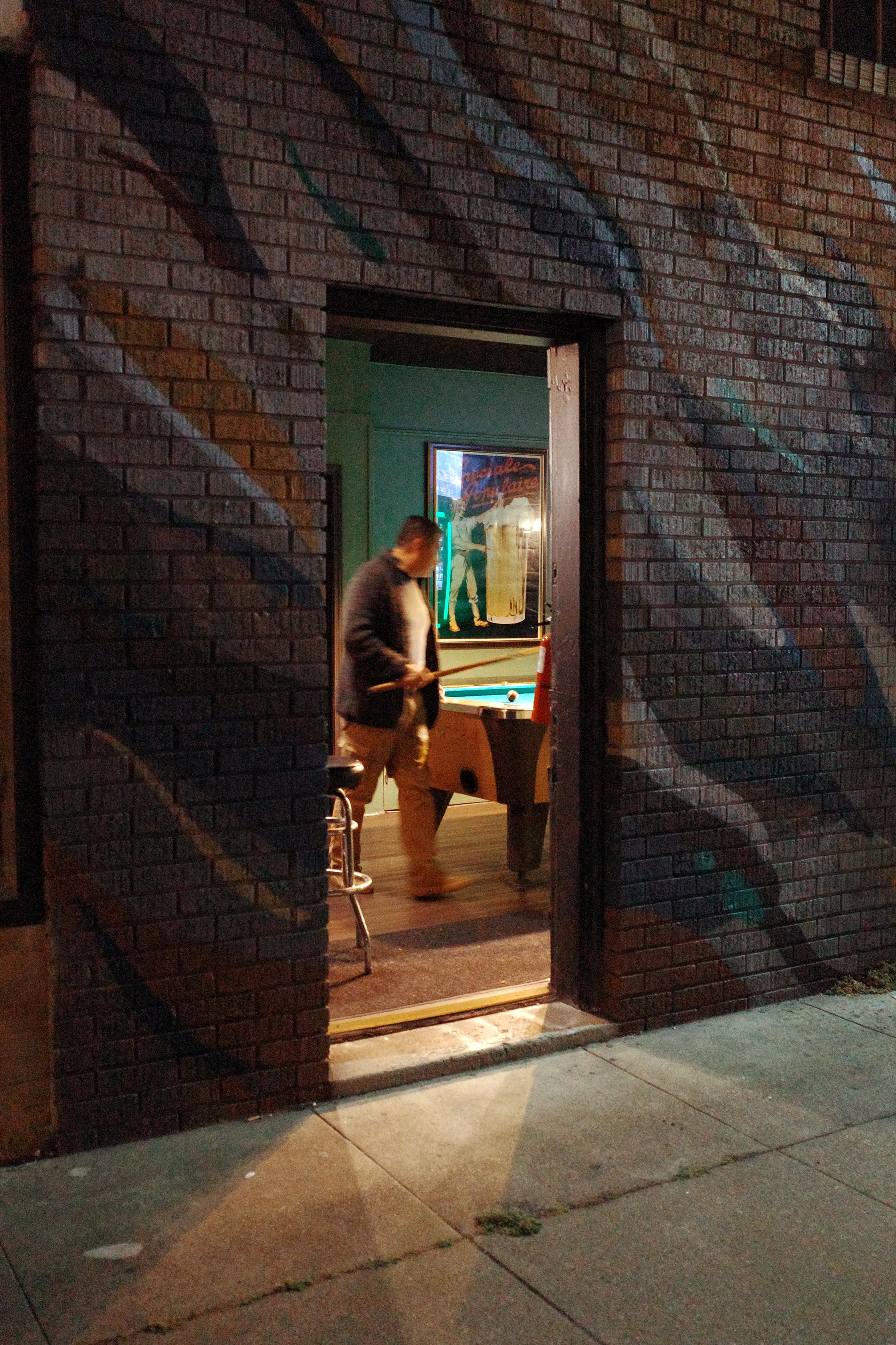 A photo shot from the sidewalk looking at a man playing pool inside of a bar